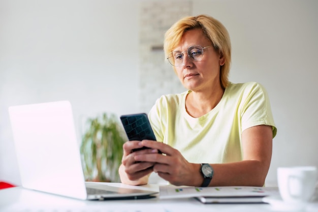 Photo image latérale d'une femme d'âge moyen avec un smartphone à la main lors d'un achat en ligne surfant sur internet une femme portant des lunettes en train de discuter au téléphone pendant son travail au bureau à domicile