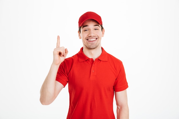 Image d'un livreur optimiste en t-shirt rouge et casquette souriant et pointant le doigt vers le haut sur le texte ou le produit de fond, isolé sur un espace blanc