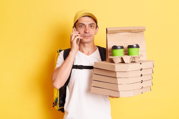 Image d'un livreur sérieux avec un sac à dos thermo en t-shirt blanc et une casquette debout avec une pizza un café à emporter parlant avec son client regardant la caméra isolée sur fond jaune