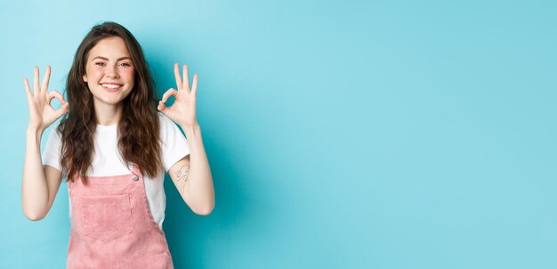 Image d'une mannequin brune souriante disant oui montrant des signes corrects d'approbation d'accord ou louant un bon choix recommandant un produit debout sur fond bleu