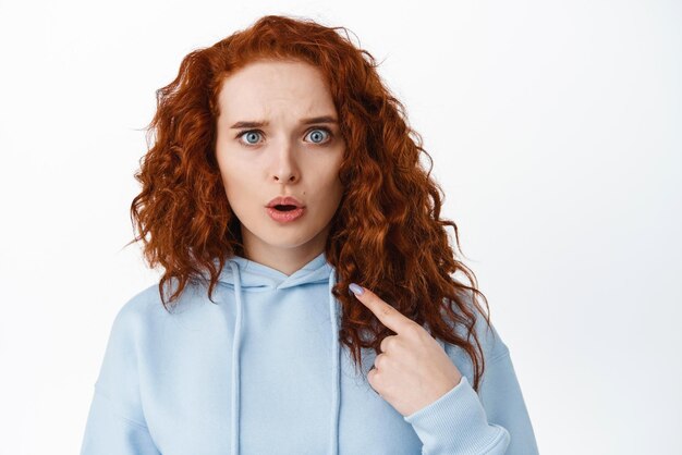 Image d'une mannequin rousse confuse et choquée pointant vers elle-même et ayant l'air inquiète d'être accusée ou blâmée debout en sweat à capuche sur fond blanc