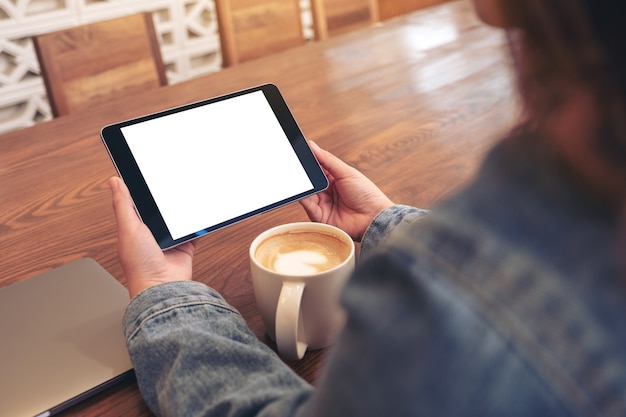 Image de maquette d'une femme tenant un tablet pc noir avec écran blanc vide horizontalement avec tasse de café et ordinateur portable sur table en bois
