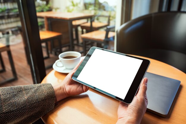 Image maquette d'une femme tenant une tablette noire avec un écran blanc vierge avec un ordinateur portable et une tasse à café sur une table en bois