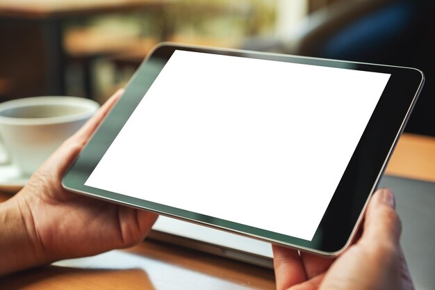 Image maquette d'une femme tenant une tablette noire avec un écran blanc vierge avec un ordinateur portable et une tasse à café sur une table en bois