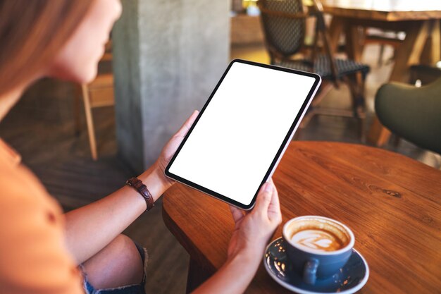 Image maquette d'une femme tenant une tablette numérique avec un écran de bureau blanc vierge avec une tasse de café sur une table en bois