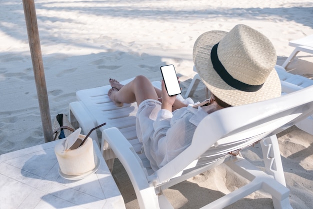 Image de maquette d'une femme tenant un téléphone mobile blanc avec écran de bureau vide tout en fixant sur une chaise de plage sur la plage