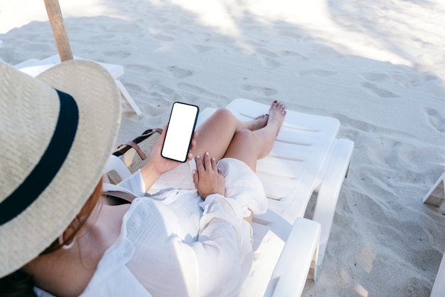 Image maquette d'une femme tenant un téléphone portable blanc avec un écran de bureau vierge tout en s'allongeant sur une chaise de plage sur la plage