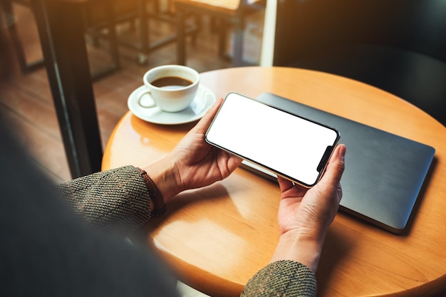 Image maquette d'une femme tenant un téléphone portable avec un écran de bureau blanc vierge avec un ordinateur portable et une tasse à café sur la table