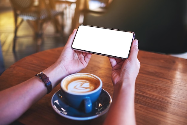 Image maquette d'une femme tenant un téléphone portable avec un écran de bureau blanc vierge avec une tasse de café sur la table