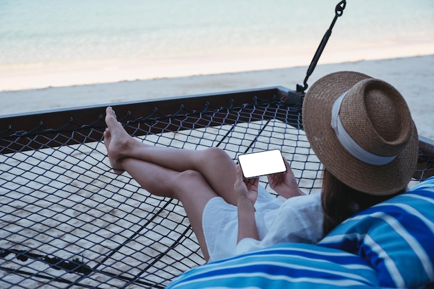 Image maquette d'une femme tenant un téléphone portable avec un écran de bureau vierge en position allongée sur un hamac sur la plage