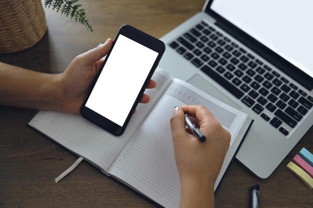 Image maquette d'une main de femme tenant un smartphone à écran de bureau blanc vierge avec un ordinateur portable sur une table tout en écrivant son idée sur un ordinateur portable. Achats en ligne, Marketing numérique sur téléphone mobile