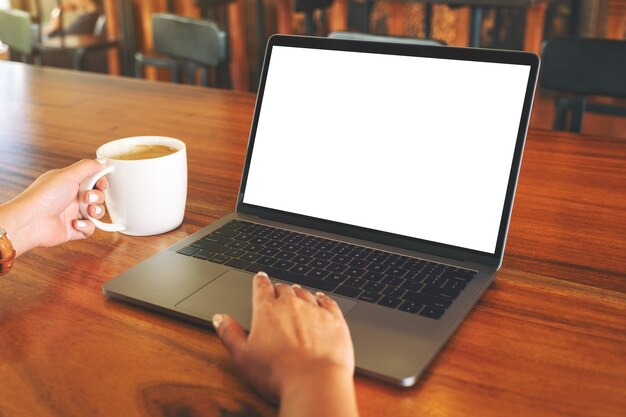 Image maquette de la main d'une femme utilisant et touchant le pavé tactile d'un ordinateur portable avec un écran de bureau blanc vierge tout en buvant du café sur une table en bois