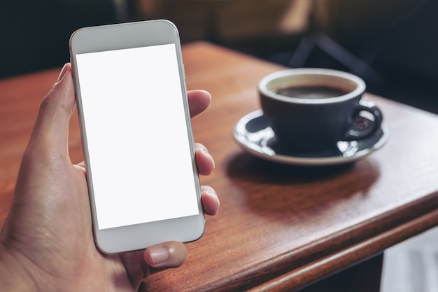 Image maquette de main tenant un téléphone mobile blanc avec écran de bureau vide avec une tasse de café sur une table en bois au café