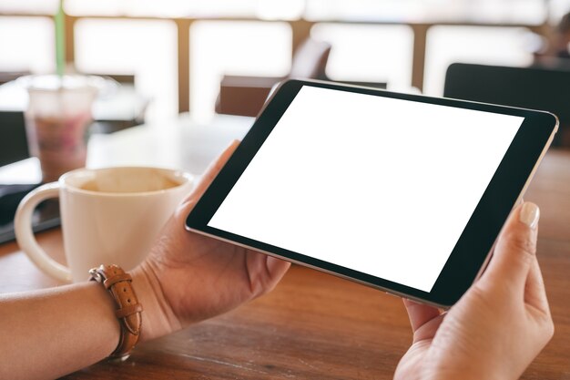 Image de maquette de mains tenant un tablet pc noir avec écran blanc vierge horizontalement avec une tasse de café sur une table en bois