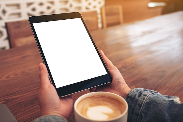 Image de maquette de mains tenant un tablet pc noir avec écran blanc vierge avec une tasse de café sur une table en bois