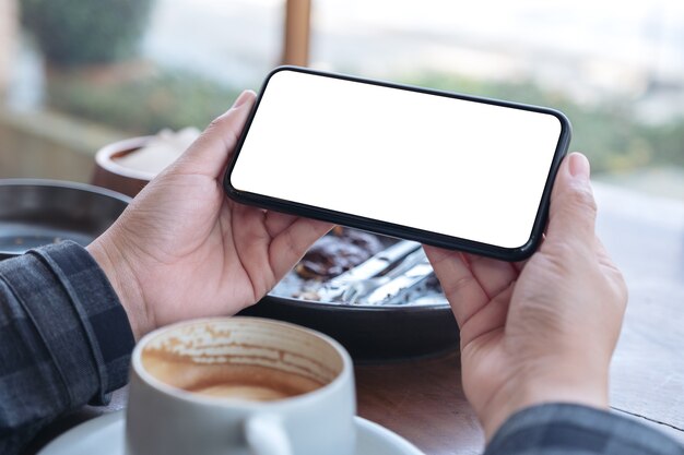 Image de maquette de mains tenant un téléphone mobile noir avec écran de bureau vide horizontalement avec une tasse de café sur la table