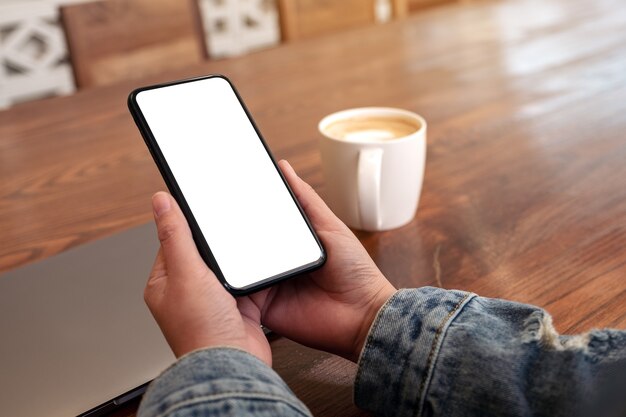 Image de maquette de mains tenant un téléphone mobile noir avec écran de bureau vide avec ordinateur portable et tasse de café sur la table