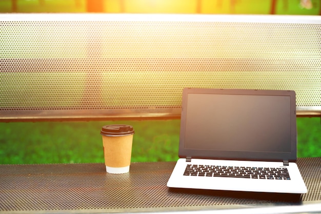 Image maquette d'un ordinateur portable avec écran noir vierge et tasse à café sur un banc en métal dans un parc extérieur naturel