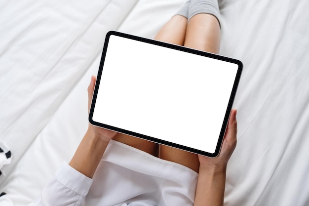 Image de maquette vue de dessus d'une femme tenant une tablette noire avec un écran blanc de bureau vierge en position allongée sur un lit blanc confortable à la maison