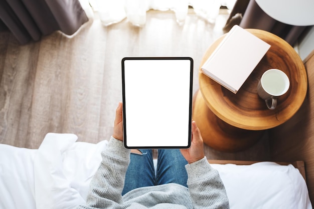 Image de maquette vue de dessus d'une femme tenant une tablette noire avec un écran blanc de bureau vierge tout en étant assise sur un lit blanc confortable à la maison