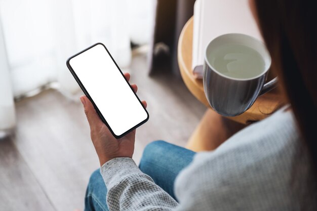 Image de maquette vue de dessus d'une femme tenant un téléphone portable avec un écran de bureau vierge tout en buvant de l'eau chaude dans la chambre