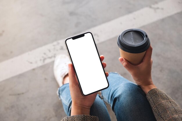 Image de maquette vue de dessus d'une femme tenant un téléphone portable noir avec un écran blanc vierge tout en buvant du café