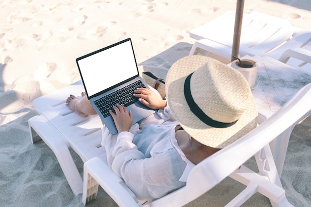 Image de maquette vue de dessus d'une femme tenant et utilisant un ordinateur portable avec écran de bureau vide tout en fixant sur une chaise de plage sur la plage