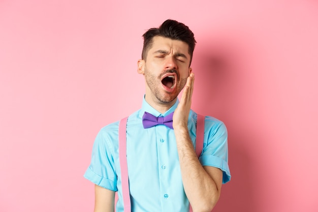 Image d'un mec fatigué en noeud papillon bâillant avec la bouche ouverte et les yeux fermés, debout somnolent, se sentant fatigué, debout sur fond rose.