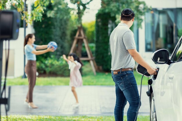 Image de mise au point d'un homme progressiste chargeant une voiture électrique avec son arrière-plan familial flou