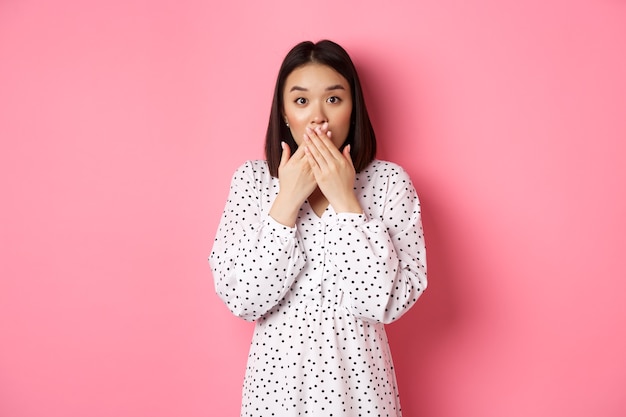 Image d'un modèle féminin asiatique surpris haletant, couvrant la bouche et regardant la caméra, debout étonné sur fond rose.