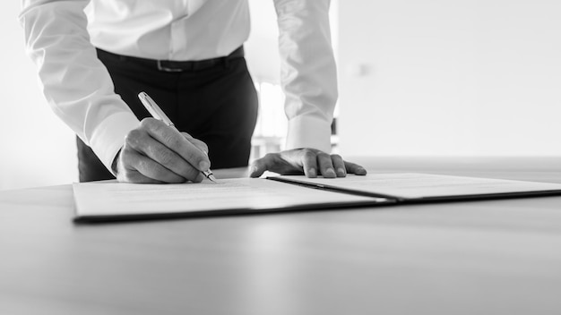 Image en niveaux de gris d'un homme d'affaires debout à son bureau signant un contrat ou un document juridique