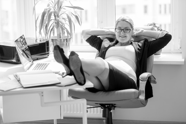 Image en noir et blanc d'une femme d'affaires enceinte souriante assise sur une chaise et tenant des jambes sur une table de bureau