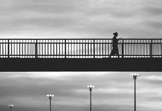 Image en noir et blanc d'une femme silhouette utilisant un smartphone tout en marchant sur un viaduc au coucher du soleil