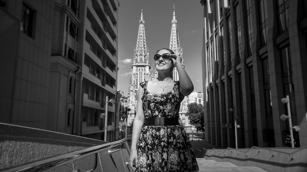 Image en noir et blanc d'une jeune femme souriante à lunettes de soleil marchant dans la rue avec un bâtiment moderne et des cathédrales anciennes