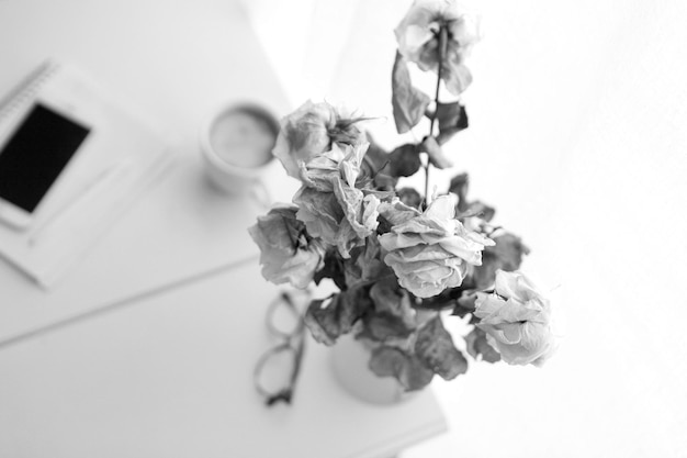 Image en noir et blanc de vieilles roses à la table de travail comme concept de procrastination ou de retard ou d'épuisement. style vintage - lunettes, carnet de notes avec stylo crayon et tasse à café sur la table