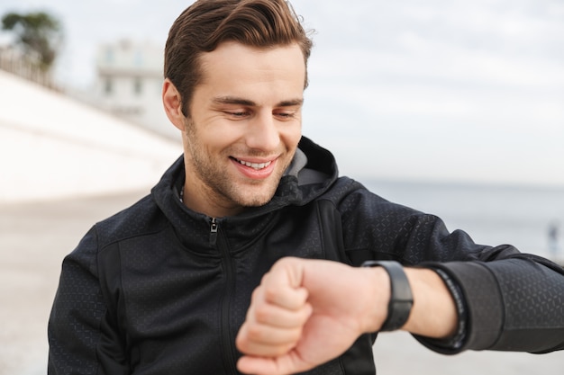 Image of smiling guy 30s en vêtements de sport noir regardant montre-bracelet, assis sur la promenade au bord de la mer