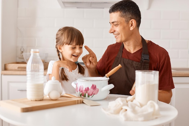 Image d'un père brun heureux et excité portant un t-shirt bordeaux cuisant avec sa fille dans la cuisine alors qu'il était assis à table, le père a enduit le nez de l'enfant de farine.