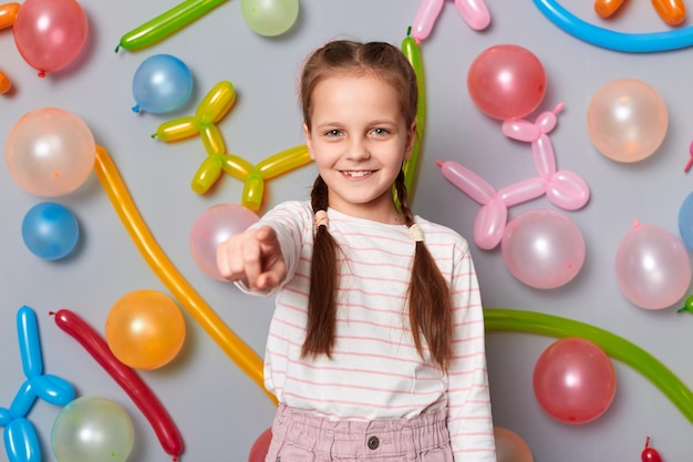 Image d'une petite fille amicale et satisfaite souriante avec des tresses portant des vêtements décontractés pointant vers la caméra invitant à sa fête posant isolée sur fond gris avec des ballons