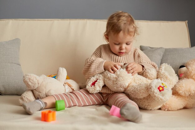 Image d'une petite fille attentive et concentrée en pull beige assise sur la toux avec un gros lapin en peluche essayant de trouver le nez en regardant une peluche avec une expression concentrée