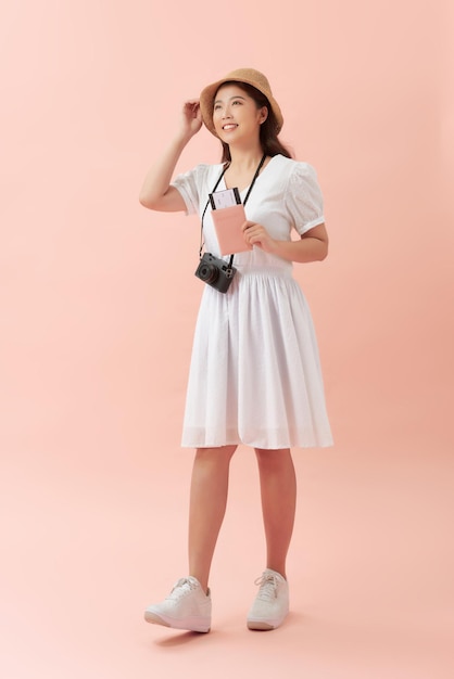 Image pleine longueur d'une femme asiatique souriante au chapeau avec un appareil photo rétro tenant un passeport avec des billets et regardant l'appareil photo sur fond rose