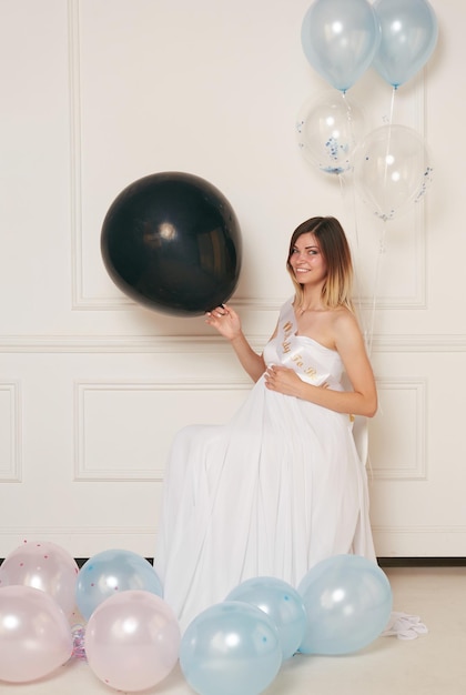 Une image pleine longueur d'une jeune femme enceinte souriante dans une robe blanche étreint le mur néoclassique blanc isolé du ventre