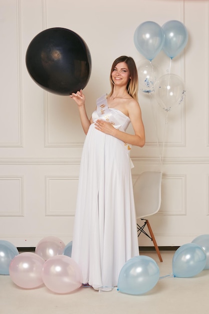 Image pleine longueur d'une jeune femme enceinte vêtue d'une robe blanche étreignant le ventre tenant un ballon noir