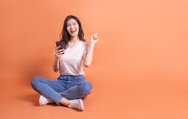 Image pleine longueur d'une jeune fille asiatique à l'aide d'un smartphone sur fond orange