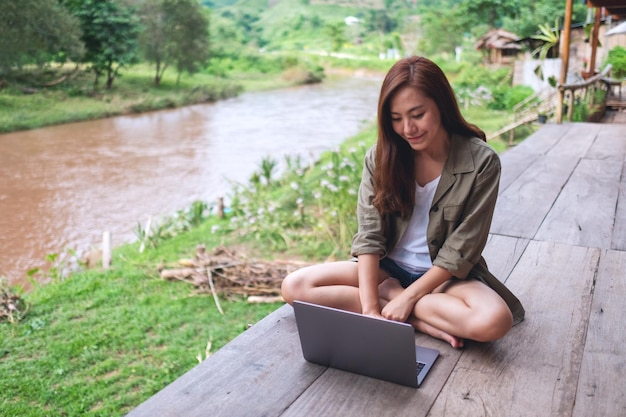 Image portrait d'une belle femme asiatique travaillant sur un ordinateur portable alors qu'elle était assise au bord de la rivière avec des montagnes et un fond de nature