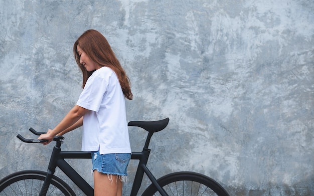 Image de portrait d'une belle jeune femme asiatique portant un t-shirt blanc et un pantalon court en jean avec un vélo et un fond de mur en béton