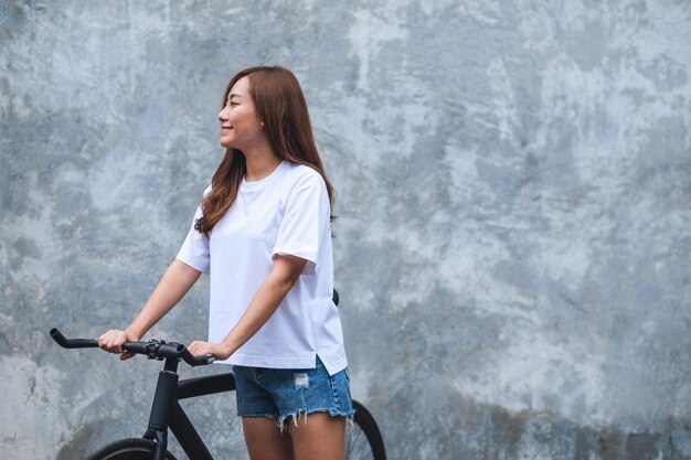 Image de portrait d'une belle jeune femme asiatique portant un t-shirt blanc et un pantalon court en jean avec un vélo et un fond de mur en béton