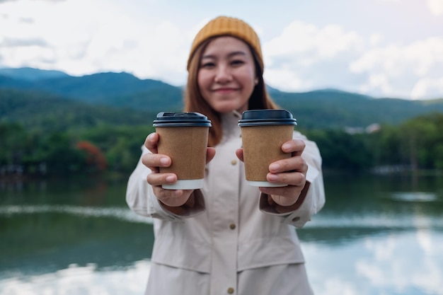 Image de portrait d'une jeune femme tenant et donnant deux tasses de café tout en voyageant dans les montagnes et le lac