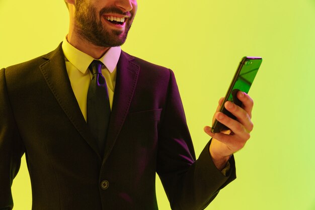 Image recadrée d'un beau jeune homme d'affaires heureux portant un costume formel debout isolé sur un mur jaune, regardant un téléphone portable