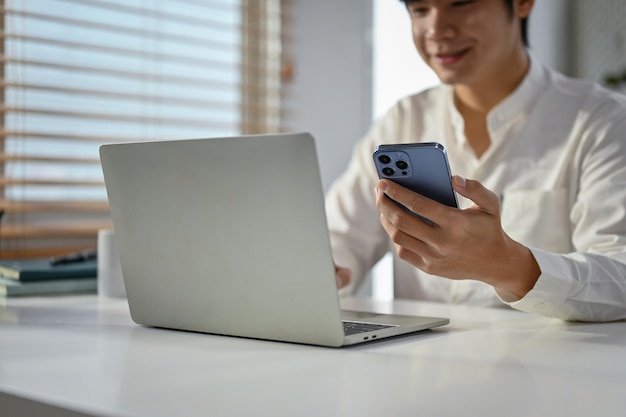 Photo image recadrée d'un bel homme d'affaires asiatique utilisant son téléphone et son ordinateur portable à son bureau
