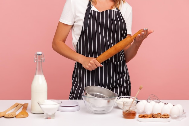 Image recadrée d'un boulanger en tablier rayé, tenant un rouleau à pâtisserie dans les mains, entouré d'ingrédients pour faire de la pâtisserie, cuisiner des gâteaux. Tourné en studio intérieur isolé sur fond rose.